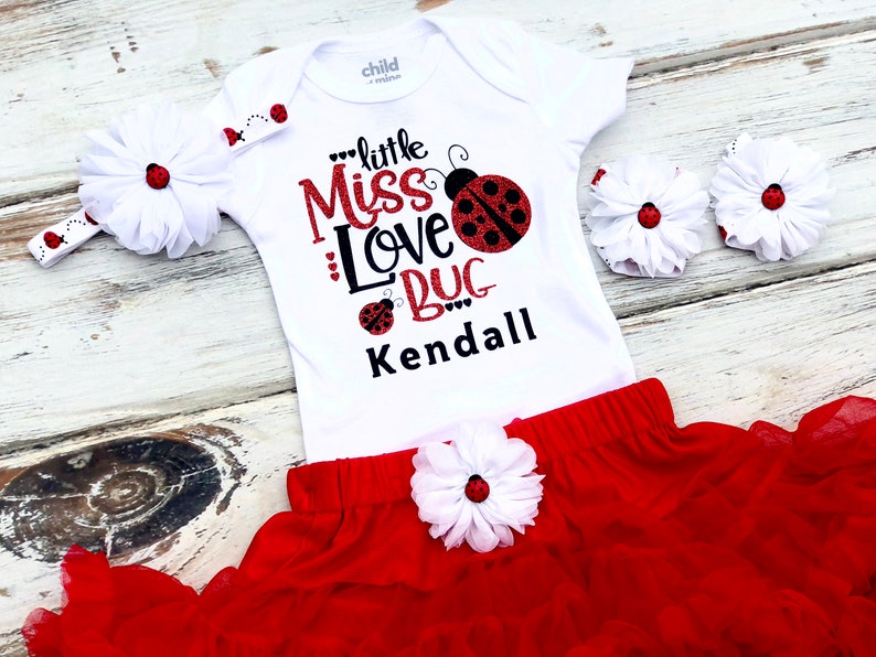May include: A white bodysuit with a red and black ladybug design and the text "little Miss Love Bug Kendall". The outfit includes a red tulle skirt with a white flower and a white headband with a red ladybug.