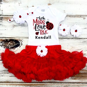 May include: A white bodysuit with the text "little Miss Love Bug Kendall" and a red ladybug graphic. A red tulle skirt with a white flower and a white headband with a red ladybug are included.