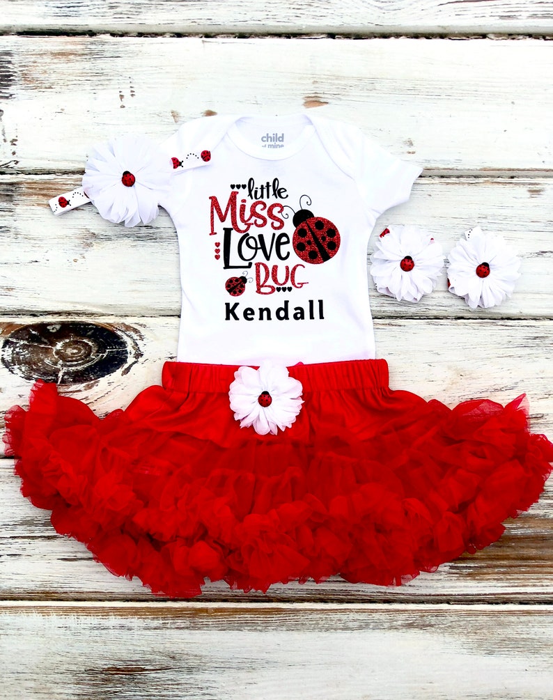May include: A white bodysuit with the text "little Miss Love Bug Kendall" and a red ladybug graphic. A red tulle skirt with a white flower and a white headband with a red ladybug are included.