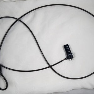 May include: A black cable lock with a combination lock. The lock has a black plastic body and a silver metal keyhole. The cable is coiled and is about 1 meter long.