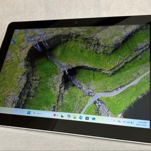 May include: A black tablet with a bright screen displaying a scenic landscape of green hills, a road, and a waterfall. The tablet has a black bezel and a silver frame. The screen shows the Windows 10 interface.
