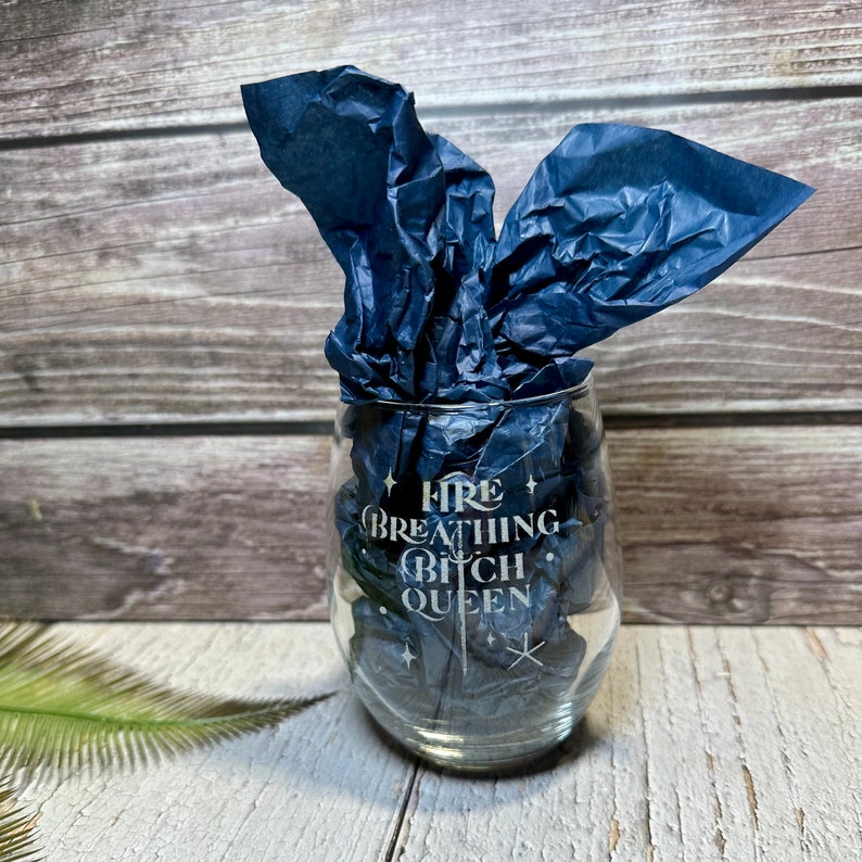May include: A clear glass wine glass with the text "Fire Breathing Bitch Queen" etched on the side. The glass is sitting on a wooden surface and is partially covered with blue tissue paper.