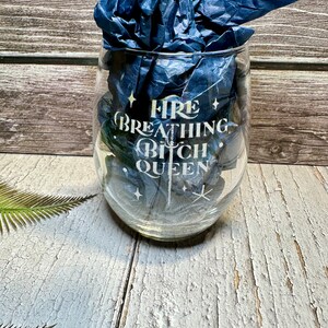 May include: A clear glass wine glass with the text "Fire Breathing Bitch Queen" etched on the side. The glass is sitting on a wooden surface.