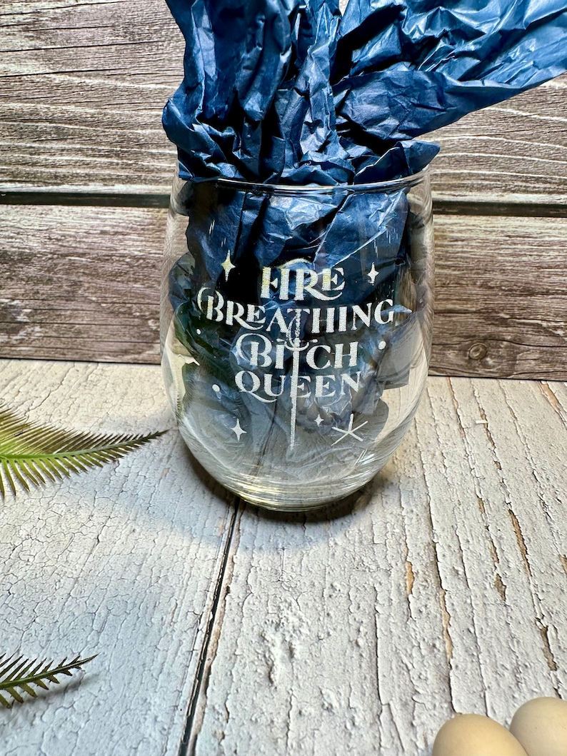 May include: A clear glass wine glass with the text "Fire Breathing Bitch Queen" etched on the side. The glass is sitting on a wooden surface.