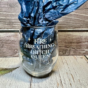 May include: A clear glass wine glass with a white design that says "Fire Breathing Bitch Queen" with a sword and stars.