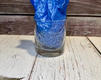 Könnte beinhalten: Ein klares Glas mit einem weißen, geätzten Logo, das The Desperado Club liest, mit dem kleineren Text So Fun It Hurts darunter. Das Glas ist mit zerknittertem blauem Seidenpapier gefüllt, was einen lebendigen Kontrast zum klaren Glas erzeugt.