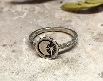 May include: A silver ring with a crescent moon and star design. The ring is on a textured, light gray surface.