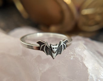May include: A silver ring with a bat design. The bat is facing forward with its wings spread out. The ring is on a pink and white background.