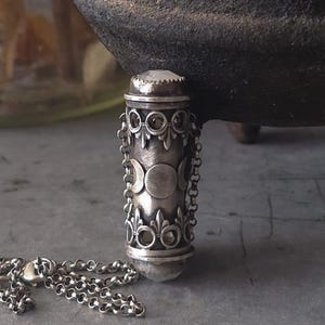 May include: A silver-colored pendant necklace with a cylindrical charm. The charm features a series of crescent moon designs and decorative filigree. A small chain is attached to the charm. The pendant has a small, clear stone on top.