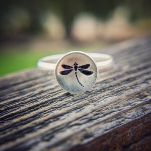 May include: A silver ring with a round, textured top featuring a black dragonfly design.