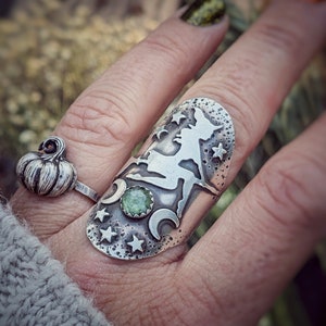 May include: A silver ring featuring a witch silhouette, stars, and crescent moons. A green gemstone is set in the centre of the ring.