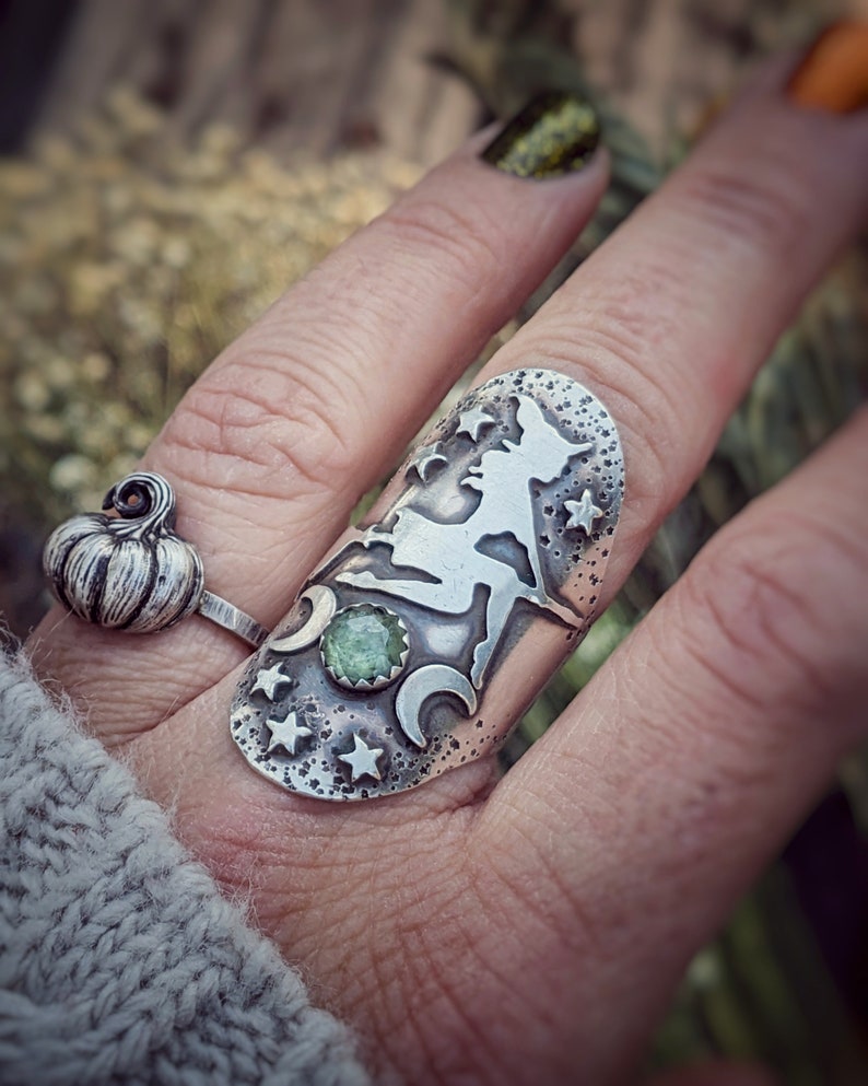 May include: A silver ring featuring a witch silhouette, stars, and crescent moons. A green gemstone is set in the centre of the ring.