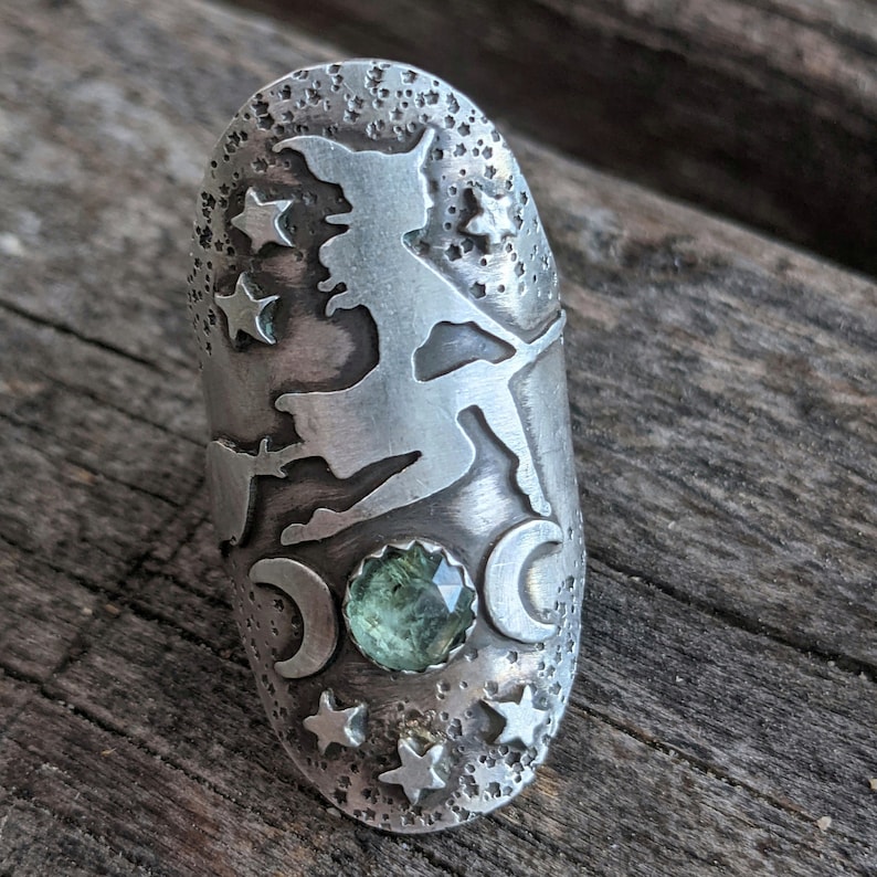 May include: A silver ring featuring a witch silhouette, stars, and crescent moons. A green gemstone is set in the centre.