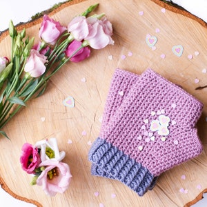 May include: A pair of pink and gray crocheted fingerless gloves with a butterfly design on the back. The gloves are on a wooden surface with pink and white hearts scattered around them.