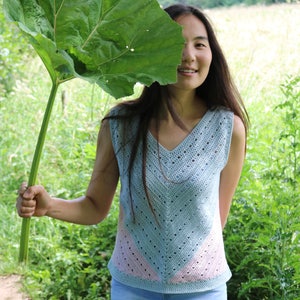 May include: A woman wearing a light blue crochet top with a pink and blue pattern. She is holding a large green leaf in front of her face.