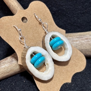 Può includere: Un paio di orecchini pendenti con fette circolari di corno con un esterno marrone ruvido e un interno bianco. Ogni orecchino contiene due perline a disco blu turchese brillante impilate, sospese da un gancio in metallo color argento.