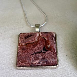 May include: A square pendant necklace featuring a reddish-brown stone with a unique, cracked pattern. The stone is set in a silver-colored metal frame and hangs from a delicate silver chain. The pendant is approximately 1.5 inches square.