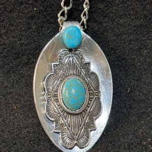 May include: Silver-toned pendant necklace featuring a turquoise stone and decorative embellishments. The pendant is oval-shaped with a detailed design around a central turquoise stone. The necklace has a silver chain.