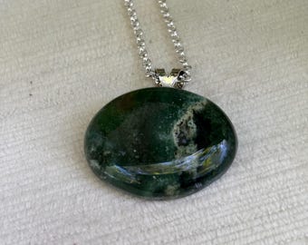 May include: A green moss agate pendant necklace with a silver chain. The oval-shaped stone has a polished surface with dark green and brown inclusions. The silver chain is delicate and attached to the stone with a small silver bail.