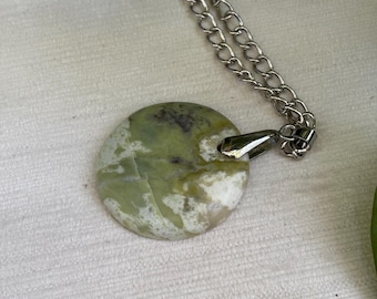 May include: A round pendant necklace featuring a green and white stone with a silver-colored chain. The stone has a marbled appearance with variations in color. The chain is a simple link style, and the pendant is attached with a silver-toned bail.