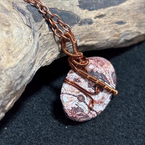 May include: A handcrafted necklace featuring a marbled stone pendant in shades of red, white, and brown. The stone is wrapped in copper wire and suspended from a copper chain. The necklace is displayed on a piece of driftwood.