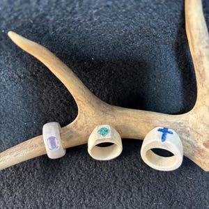 May include: Three handcrafted rings made from a light-colored material, possibly bone or antler, each with unique designs. One ring features a rectangular purple stone, another has a turquoise inlay, and the third has a blue cross design.