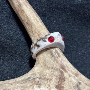 Könnte beinhalten: Ein Ring aus hellfarbigem, texturiertem Geweihmaterial mit einer glatten, polierten Oberfläche. Das Band ist mit zwei kleinen, kreisförmigen, leuchtend roten Edelsteinen besetzt, die bündig in das Material eingelassen sind.