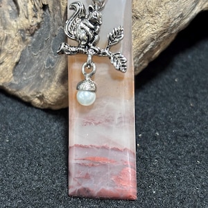 Puede incluir: Colgante rectangular con una ardilla plateada sobre una rama, sosteniendo una perla. El colgante tiene una piedra rosa y blanca con vetas rojas. El colgante mide aproximadamente 5 cm de largo.