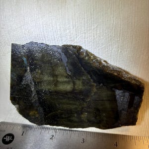May include: A rough-cut labradorite gemstone with dark green, blue, and black colours. A metal ruler with inch markings is placed next to the stone for scale. The stone measures approximately 10 cm in length.