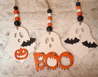 May include: Three wooden Halloween ornaments with orange and black paint. The ornaments are shaped like ghosts and bats. One ghost has a pumpkin face, another has the word "BOO" written on it, and the third has two bats on it.