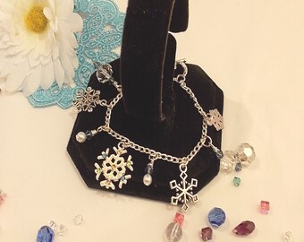 May include: Silver-toned charm bracelet featuring snowflake charms and dangling beads. The bracelet includes clear, blue, and purple crystal beads, and is displayed on a black velvet stand. A white flower and lace are in the background.