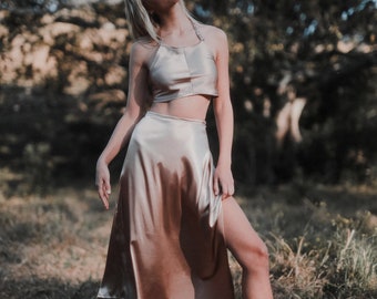 May include: A woman wearing a gold satin crop top and matching skirt with a slit. She is standing in a field of tall grass.