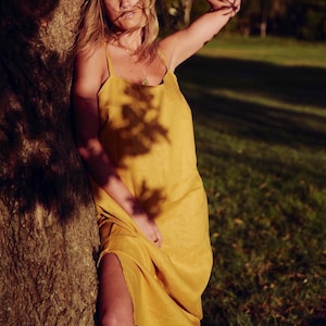 Puede incluir: Una mujer con un vestido de lino amarillo se encuentra de pie junto a un &aacute;rbol. El vestido tiene un dise&ntilde;o sencillo, sin mangas y un ajuste holgado. El cabello de la mujer es largo y suelto, y est&aacute; mirando hacia un lado.