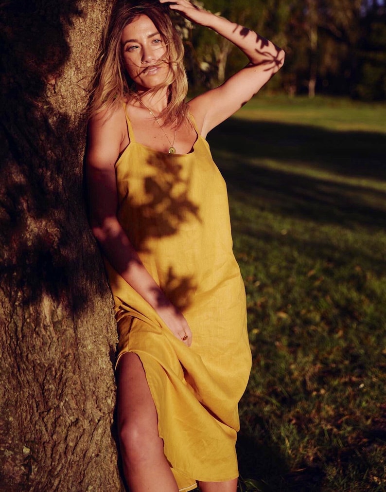 Puede incluir: Una mujer con un vestido de lino amarillo se encuentra de pie junto a un &aacute;rbol. El vestido tiene un dise&ntilde;o sencillo, sin mangas y un ajuste holgado. El cabello de la mujer es largo y suelto, y est&aacute; mirando hacia un lado.