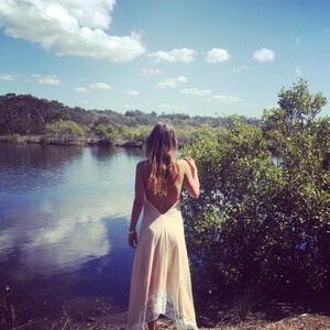 Op de afbeelding: Een vrouw in een lange, lichtroze jurk met kant staat op een houten bankje met uitzicht op een waterpartij. Het water is kalm en reflecteert de blauwe lucht en witte wolken. De vrouw heeft lang, donker haar en kijkt uit over het water.