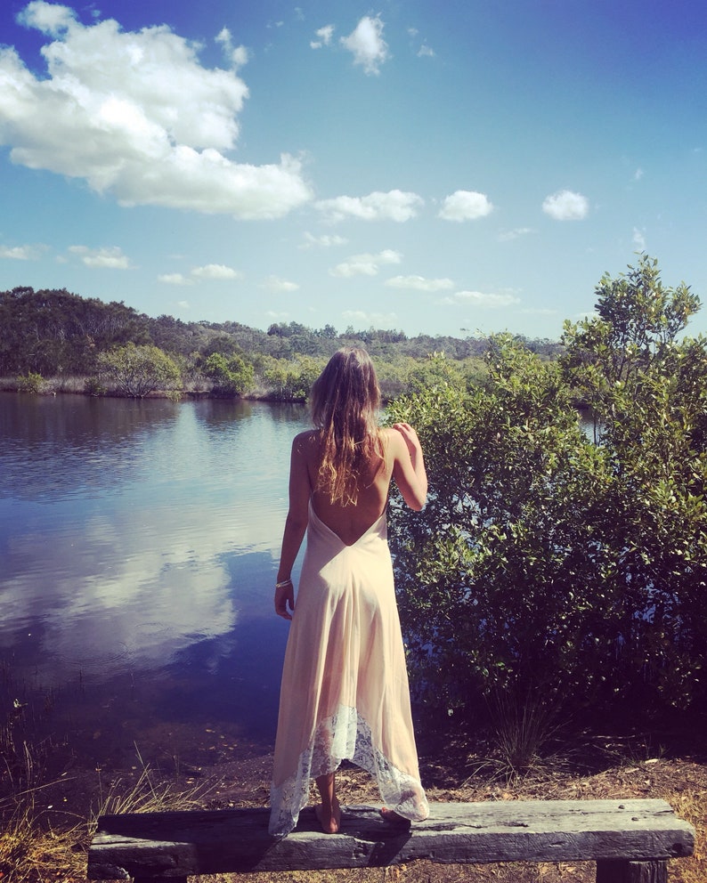 Op de afbeelding: Een vrouw in een lange, lichtroze jurk met kant staat op een houten bankje met uitzicht op een waterpartij. Het water is kalm en reflecteert de blauwe lucht en witte wolken. De vrouw heeft lang, donker haar en kijkt uit over het water.