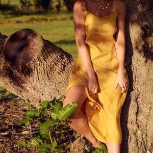Puede incluir: Una mujer con un vestido de lino amarillo se encuentra de pie frente a un tronco de &aacute;rbol. El vestido tiene un dise&ntilde;o sencillo, sin mangas y llega hasta los tobillos. Ella est&aacute; descalza y tiene el pelo largo y liso.