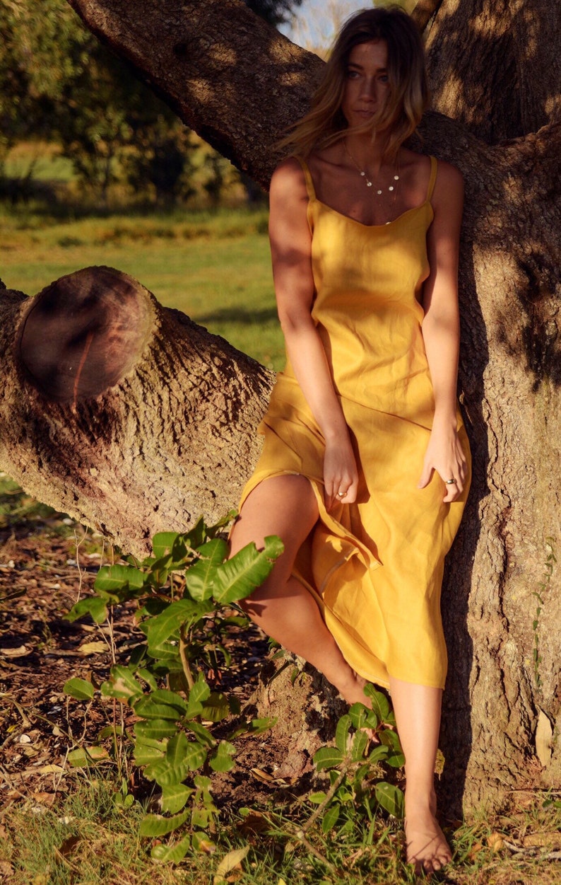 Puede incluir: Una mujer con un vestido de lino amarillo se encuentra de pie frente a un tronco de &aacute;rbol. El vestido tiene un dise&ntilde;o sencillo, sin mangas y llega hasta los tobillos. Ella est&aacute; descalza y tiene el pelo largo y liso.