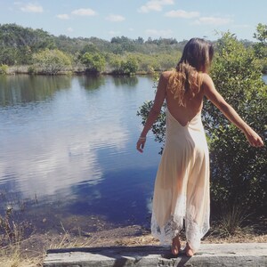 Op de afbeelding: Een vrouw in een lange, licht perzikkleurige slipdress met witte kant staat op een houten platform aan een meer. De jurk heeft een lage rug en een zwierige rok.