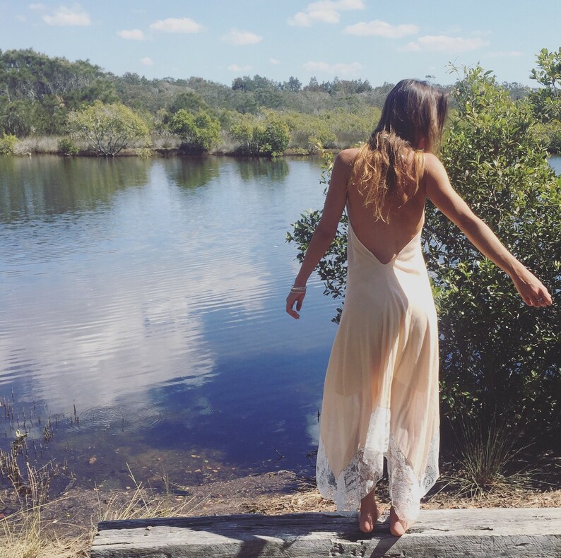 Op de afbeelding: Een vrouw in een lange, licht perzikkleurige slipdress met witte kant staat op een houten platform aan een meer. De jurk heeft een lage rug en een zwierige rok.