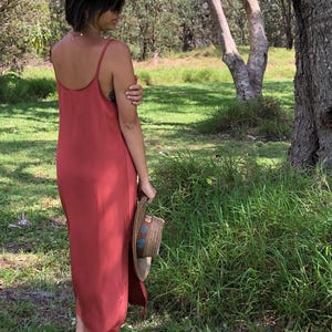 Puede incluir: Una mujer con un vestido maxi de color &oacute;xido con tirantes finos, de pie en un campo de hierba. Lleva un sombrero de paja en la mano derecha.