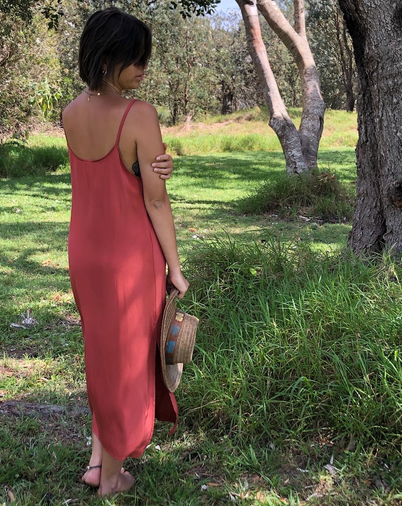 Puede incluir: Una mujer con un vestido maxi de color &oacute;xido con tirantes finos, de pie en un campo de hierba. Lleva un sombrero de paja en la mano derecha.