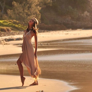 Op de afbeelding: Een vrouw in een lange, vloeiende, beige jurk met een bloemenpatroon staat op een zandstrand. Ze draagt ​​een kralenband en heeft haar hoofd naar achteren gekanteld. Het strand is leeg en het water is kalm.