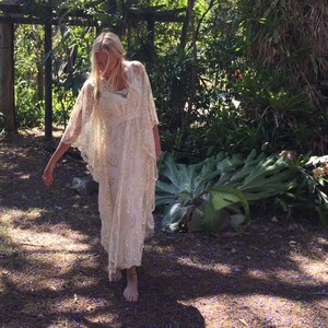 K&ouml;nnte beinhalten: Eine Frau in einem wei&szlig;en Spitzenkleid und einem wei&szlig;en Spitzenumhang steht in einem Garten mit gr&uuml;nen Pflanzen und lila Blumen auf dem Boden.