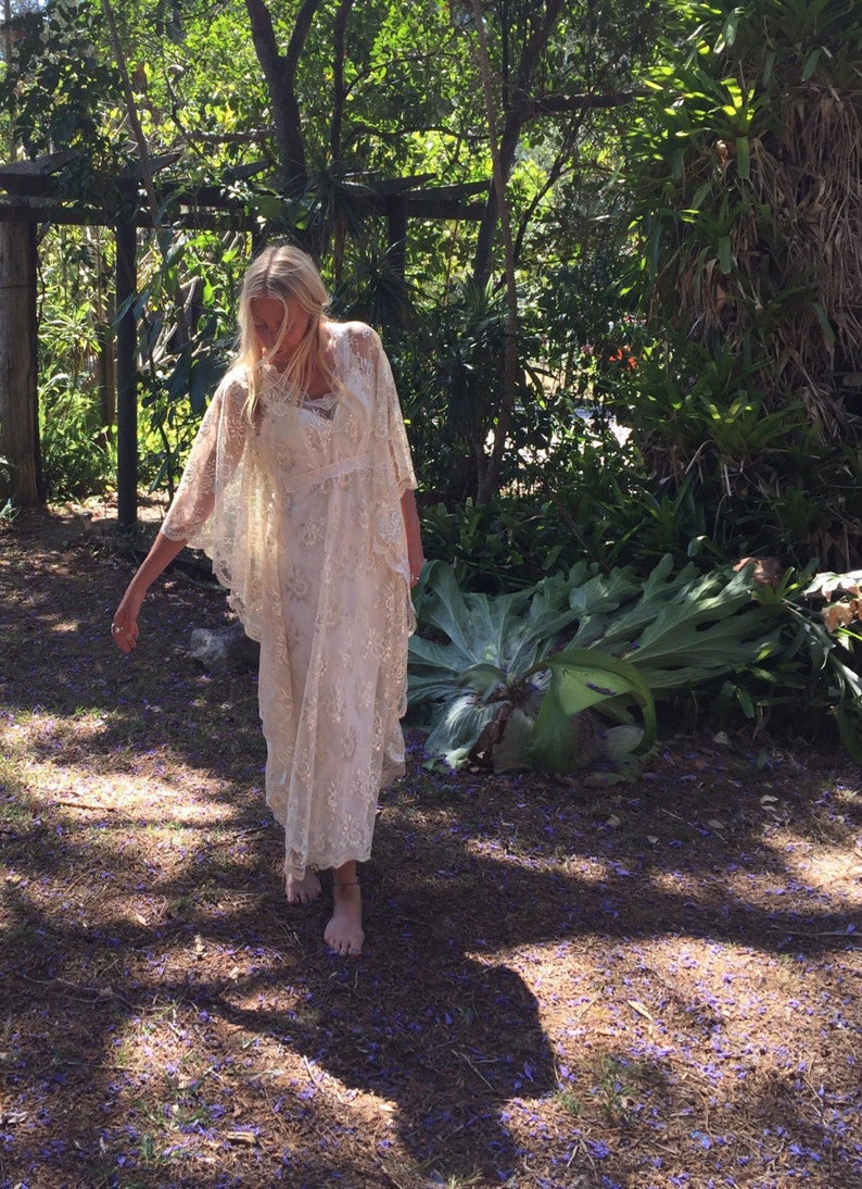 K&ouml;nnte beinhalten: Eine Frau in einem wei&szlig;en Spitzenkleid und einem wei&szlig;en Spitzenumhang steht in einem Garten mit gr&uuml;nen Pflanzen und lila Blumen auf dem Boden.