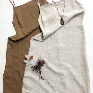 Puede incluir: Dos vestidos de lino, uno marr&oacute;n y otro beige, colgados sobre un fondo blanco. El vestido beige tiene un collar de cuero marr&oacute;n con un colgante.