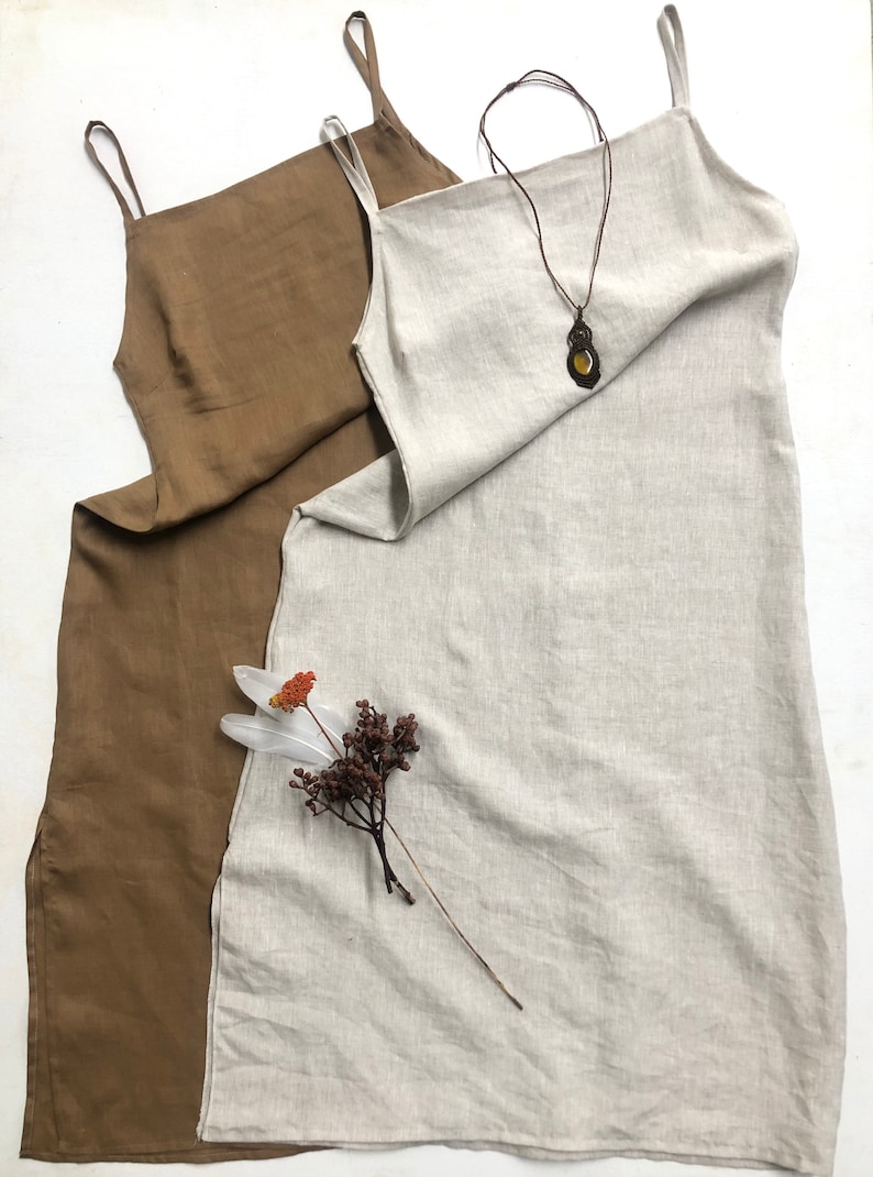 Puede incluir: Dos vestidos de lino, uno marr&oacute;n y otro beige, colgados sobre un fondo blanco. El vestido beige tiene un collar de cuero marr&oacute;n con un colgante.