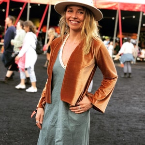 May include: A woman wearing a cream-coloured hat, a rust-coloured velvet jacket with flared sleeves, and a long, muted green dress. The background includes a circus tent and other people.