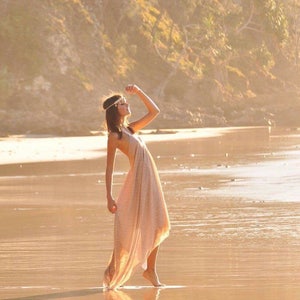 Op de afbeelding: Een vrouw in een lange, vloeiende, lichtgekleurde jurk met een kralenhoofdband staat op een zandstrand met de zon die achter haar schijnt.