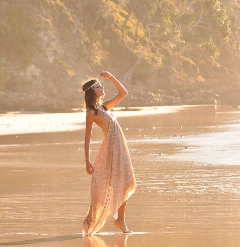 Op de afbeelding: Een vrouw in een lange, vloeiende, lichtgekleurde jurk met een kralenhoofdband staat op een zandstrand met de zon die achter haar schijnt.